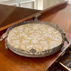 Puede incluir: Una bandeja de servicio redonda de metal con un borde plateado ornamentado y elevado que presenta figuras de aves. El centro de la bandeja está incrustado con una pieza de tela o papel con un patrón de damasco repetitivo en tonos verde oliva y crema.