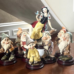 Può includere: Una collezione di statuette in ceramica raffiguranti i personaggi della fiaba di Biancaneve. Il set include Biancaneve, vestita con un abito giallo e corpetto nero, e sette nani, ognuno con abiti unici. Le statuette sono montate su basi di legno.