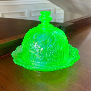 Puede incluir: Una mantequillera de vidrio verde neón vibrante con una tapa en forma de cúpula que presenta un patrón floral en relieve y un borde festoneado. La tapa está rematada con un asa escalonada y toda la pieza tiene un aspecto translúcido.
