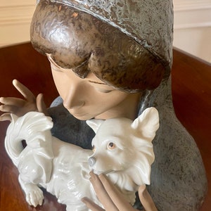 May include: A porcelain figurine of a young woman wearing a blue hooded cloak, holding a white porcelain dog. The woman's face is delicate and her eyes are closed. The dog is sitting on its hind legs and looking up at the woman.