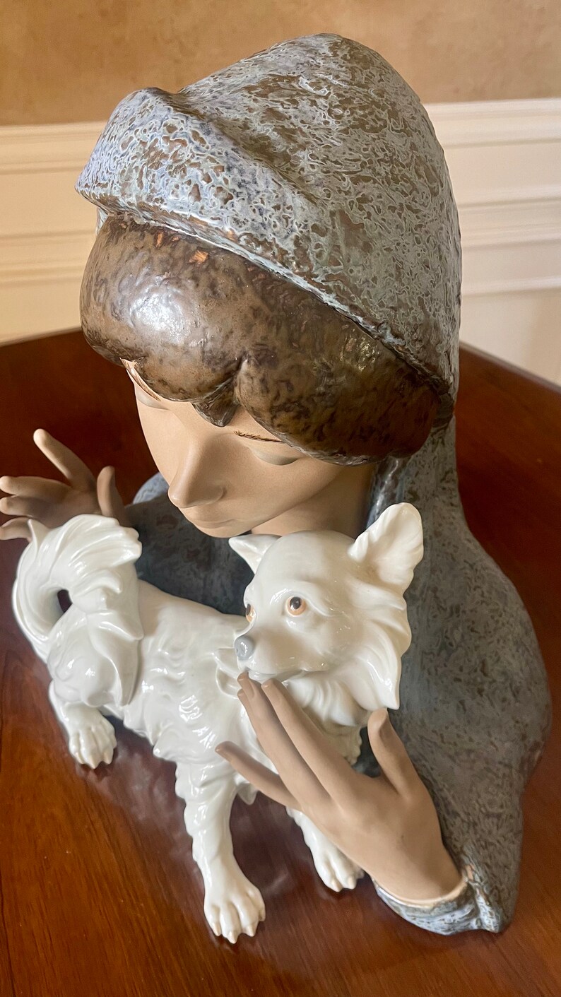 May include: A porcelain figurine of a young woman wearing a blue hooded cloak, holding a white porcelain dog. The woman's face is delicate and her eyes are closed. The dog is sitting on its hind legs and looking up at the woman.