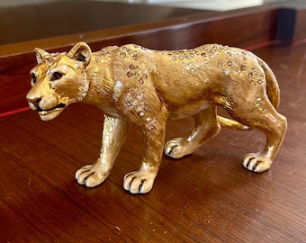 May include: A golden figurine of a lioness, adorned with sparkling embellishments. The sculpture is posed in a walking stance, showcasing intricate details. The figurine is placed on a wooden surface, adding to its decorative appeal.