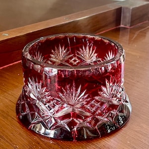 May include: A deep red crystal bowl featuring intricate cut glass patterns, including starburst motifs and geometric facets. The circular vessel has a wide, flared base and a smooth, polished rim, showcasing the vibrant ruby-colored glass construction.