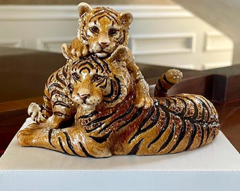 May include: A decorative figurine of a tiger and cub. The adult tiger is lying down with the cub perched on its back. The figurine is painted in shades of brown and black with gold accents. The figurine is resting on a white surface.