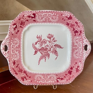 May include: A square ceramic serving platter with rounded corners and two side handles. The platter features a cream-colored center with a dark pink floral illustration and a wide, dark pink border decorated with intricate floral and scroll patterns.