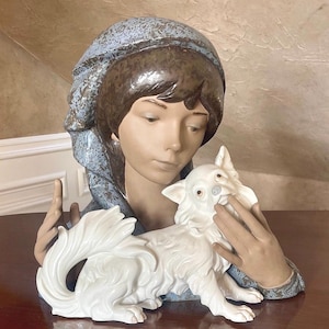 May include: A porcelain figurine of a woman with a blue and brown headscarf holding a white porcelain dog.