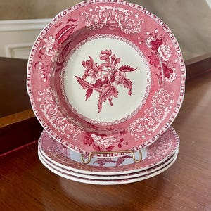 May include: A stack of three round ceramic plates featuring a deep red floral transferware pattern on a cream background. The plates have a scalloped edge and a central botanical motif surrounded by an intricate border of flowers and scrolling vines.