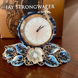 Op de afbeelding: Een gouden en blauwe tafelklok met een bij-ontwerp op de bovenkant. De wijzerplaat van de klok is wit met zwarte Romeinse cijfers en een gouden rand. De klok staat op een voet van blauwe en gouden bladeren met een witte bloem in het midden.