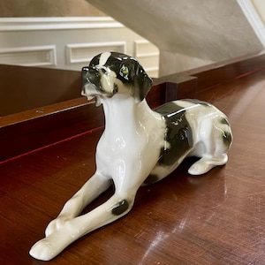 Op de afbeelding: Een porseleinen beeldje van een liggende hond. De hond heeft een witte vacht met zwarte vlekken en aftekeningen op zijn kop en rug. Het beeldje heeft groene ogen en staat op een houten oppervlak. Ongeveer 20 cm lang.