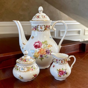 May include: A white porcelain tea set with floral designs. The set includes a teapot, sugar bowl, and creamer. The teapot has a tall, curved spout and a lid. The sugar bowl has a lid, and the creamer has a handle.