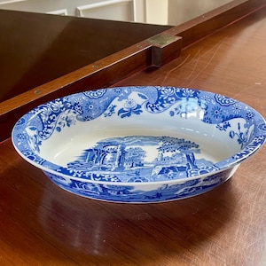 May include: An oval ceramic serving bowl featuring a white base with blue transferware patterns. The interior bottom displays a landscape scene with ruins and trees, while the rim is decorated with a repeating floral and scroll motif.