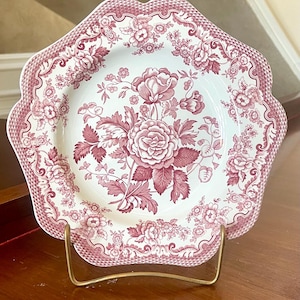 May include: A scalloped-edge ceramic plate featuring a detailed dark pink floral pattern on a cream background. The design includes a central rose motif surrounded by smaller blossoms and a decorative lattice border along the outer rim of the plate.