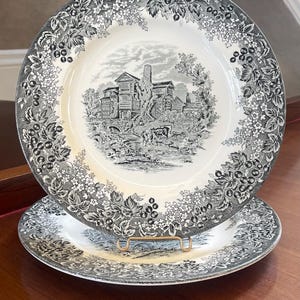 May include: Two round ceramic plates featuring a black and white transferware pattern. The centre depicts a rural landscape with a house and grazing cattle, while the wide rim is decorated with a detailed border of berries and small flowers.