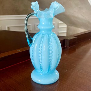 May include: A light blue glass pitcher with a ruffled rim and a beaded design. The handle is clear with a blue tint. The pitcher is sitting on a dark wooden surface.