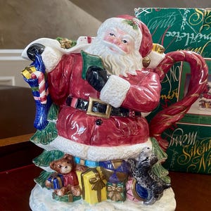 May include: A ceramic Santa Claus teapot featuring a red suit, black belt, and white beard. Santa holds a green book and a sack filled with gifts, including a blue stocking, a teddy bear, and a yellow box. The handle is a red candy cane.