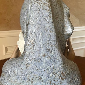 May include: A ceramic sculpture of a woman's bust, with a textured, blue and brown glaze. The woman is wearing a draped garment over her head and shoulders, and her right hand is raised to her cheek.