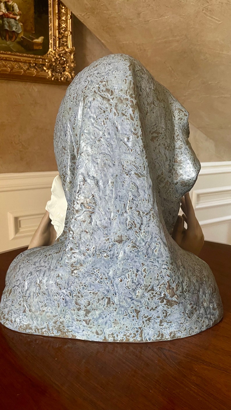 May include: A ceramic sculpture of a woman's bust, with a textured, blue and brown glaze. The woman is wearing a draped garment over her head and shoulders, and her right hand is raised to her cheek.