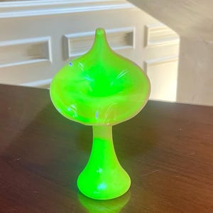Puede incluir: Un jarrón de vidrio verde neón vibrante y brillante que presenta una abertura ancha y acampanada en forma de trompeta en la parte superior y un tallo delgado y cónico que se ensancha en una base circular. El vidrio emite una luz intensa.