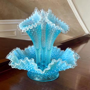 May include: A light blue glass vase with a ruffled design, featuring three central spires and a wide, scalloped base. The vase has a diamond pattern and a clear, frosted rim. It is likely vintage and sits on a dark wooden surface.