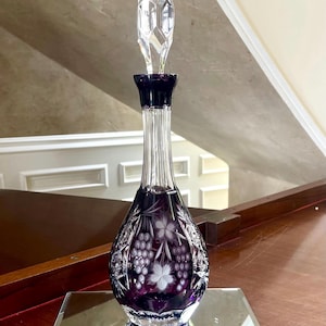 Puede incluir: Una jarra decorativa de cristal púrpura con un tapón transparente facetado. La jarra presenta un diseño de uvas y hojas y está llena de un líquido oscuro. La jarra se exhibe sobre una superficie espejada.