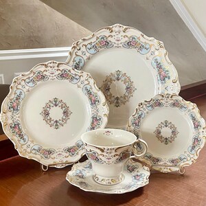 May include: A porcelain tea set featuring a large serving platter, two plates of varying sizes, and a teacup with a matching saucer. Each piece is cream-colored with scalloped edges, gold trim, and a floral pattern in pink, blue, and green.