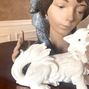 May include: A porcelain figurine of a woman with a blue and brown patterned scarf, holding a white porcelain dog. The woman has brown hair and is looking down at the dog.