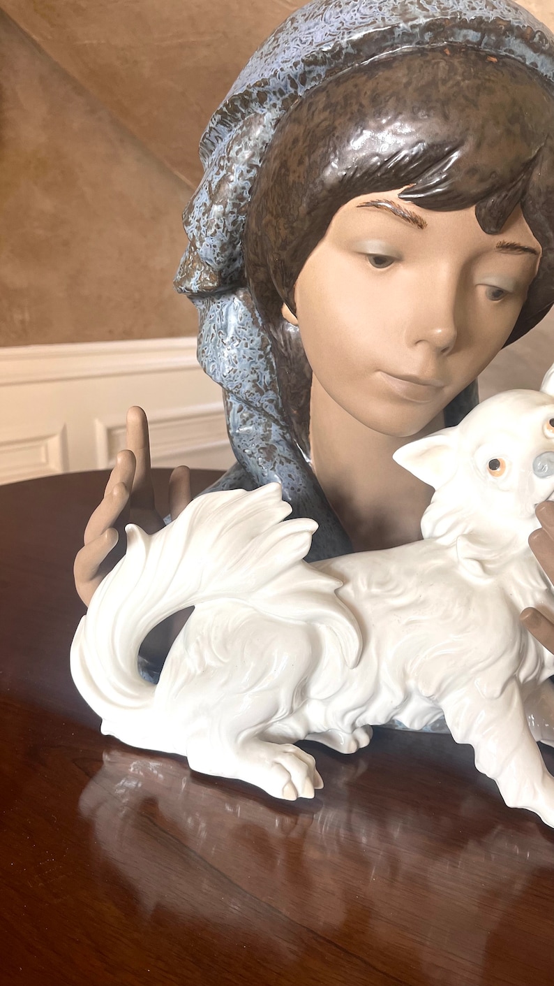 May include: A porcelain figurine of a woman with a blue and brown patterned scarf, holding a white porcelain dog. The woman has brown hair and is looking down at the dog.