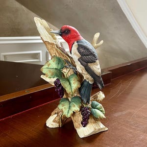 Puede incluir: Una figurita de porcelana de un pájaro carpintero de cabeza roja posado en una rama de árbol con uvas. El pájaro está pintado con detalles realistas con marcas negras y blancas en sus alas y cola.