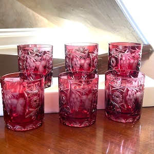 Puede incluir: Juego de seis vasos de vidrio tallado rojo rubí. Cada vaso presenta intrincados diseños florales y de estrellas. Los vasos son cilíndricos con una base gruesa, dispuestos sobre una superficie de madera.