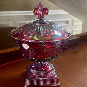Op de afbeelding: Een robijnrode, iriserende glazen bonbonnière met een deksel, met een fleur-de-lis-knop. De schaal heeft een vierkante basis en een gedetailleerd, reliëfpatroon. Het glas reflecteert meerdere kleuren.