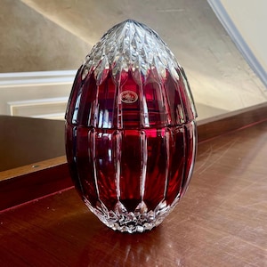 Puede incluir: Un recipiente decorativo de cristal rojo rubí en forma de huevo con tapa facetada. El recipiente tiene un diseño acanalado vertical y una base transparente facetada. Una pequeña pegatina circular es visible en la parte delantera.