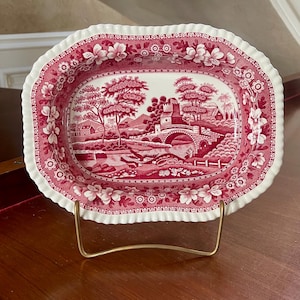 May include: An oval ceramic dish with a scalloped edge, featuring a deep red and white transferware pattern. The central design depicts a landscape with a stone bridge, trees, and buildings, while the border displays floral motifs.