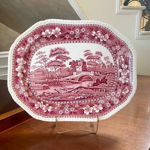 Può includere: Un piatto ovale in ceramica con un motivo a trasferimento color rosa scuro e bianco. Il centro raffigura un paesaggio con un ponte in pietra, alberi ed edifici. Il bordo smerlato è decorato con un motivo floreale e geometrico.