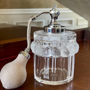 Pode incluir: Um frasco de perfume de vidro transparente com corpo cilíndrico apresentando estrias verticais e uma faixa fosca de elementos decorativos arredondados perto do topo. Inclui um mecanismo atomizador de metal prateado e uma bomba.