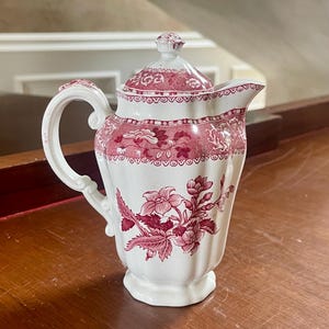 May include: A white ceramic cream jug with a lid, featuring a deep red floral transferware pattern. The vessel has a curved handle, a fluted body, and a decorative border around the top rim, showcasing detailed botanical illustrations in red ink.