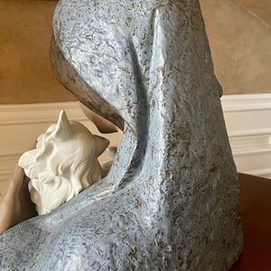 May include: A ceramic sculpture of a woman with a white dove in her arms. The woman is wearing a blue and white patterned robe and has a soft, gentle expression on her face.