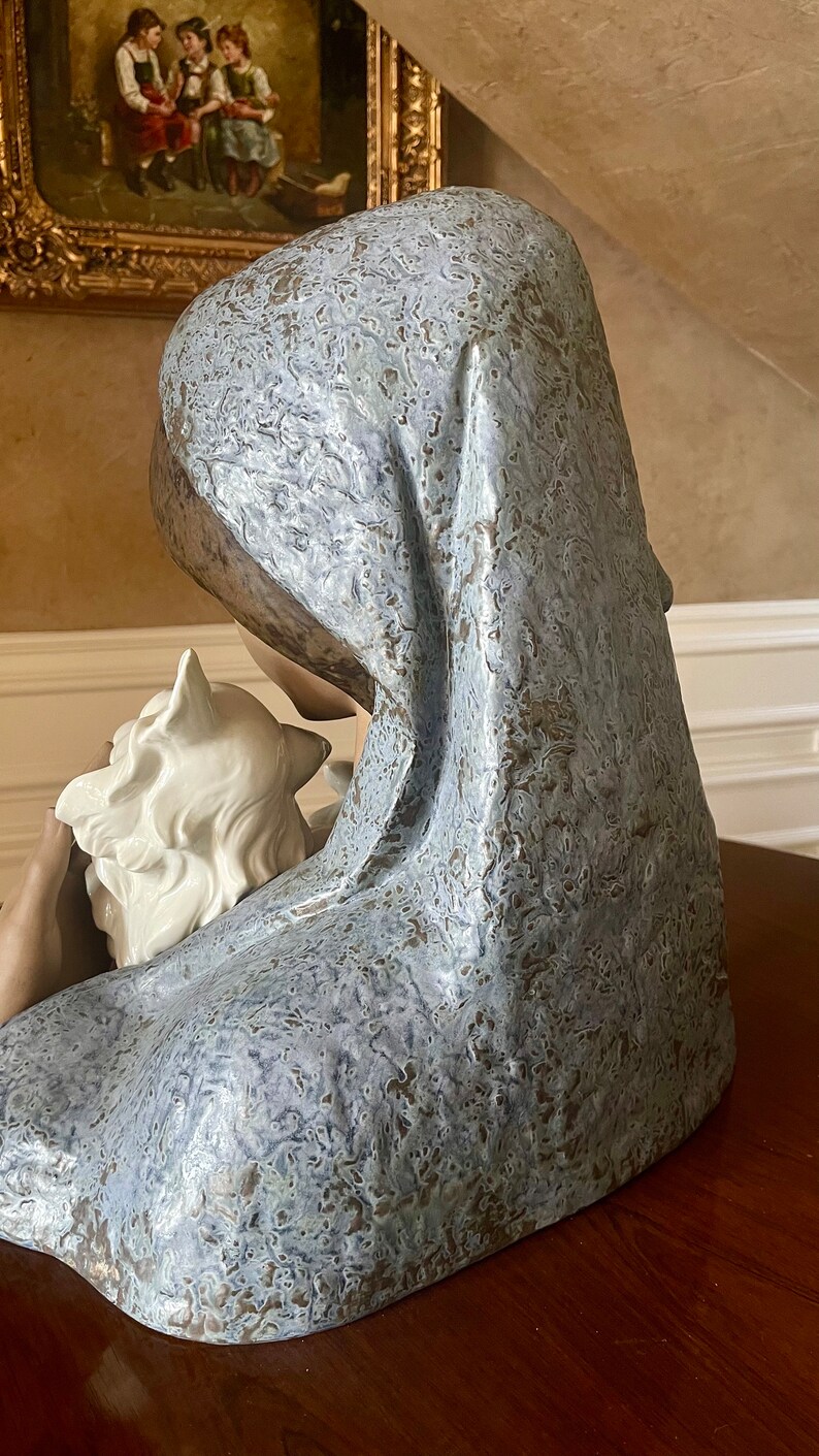 May include: A ceramic sculpture of a woman with a white dove in her arms. The woman is wearing a blue and white patterned robe and has a soft, gentle expression on her face.