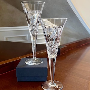 Puede incluir: Dos copas de champán de cristal transparente con un patrón de diamante y un delicado diseño de lazo en la parte superior. Las copas están sobre una caja azul oscuro.