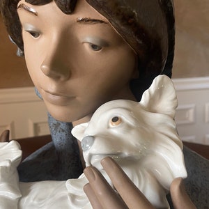 May include: A porcelain figurine of a young woman with brown hair wearing a blue and white hat, holding a white porcelain dog.