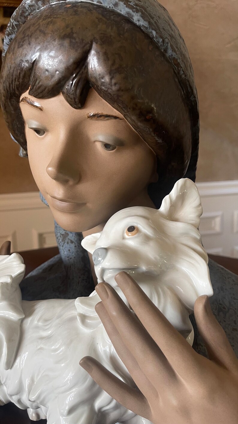 May include: A porcelain figurine of a young woman with brown hair wearing a blue and white hat, holding a white porcelain dog.