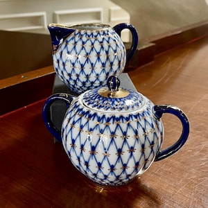 May include: A white and blue porcelain tea set with a lattice pattern and gold accents. The set includes a teapot with a lid and a creamer. The handles and rims are dark blue.