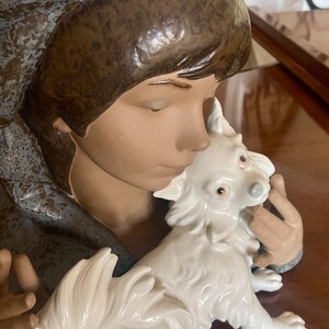 May include: A porcelain figurine of a girl with a blue and brown hooded cloak holding a white dog with a fluffy tail.