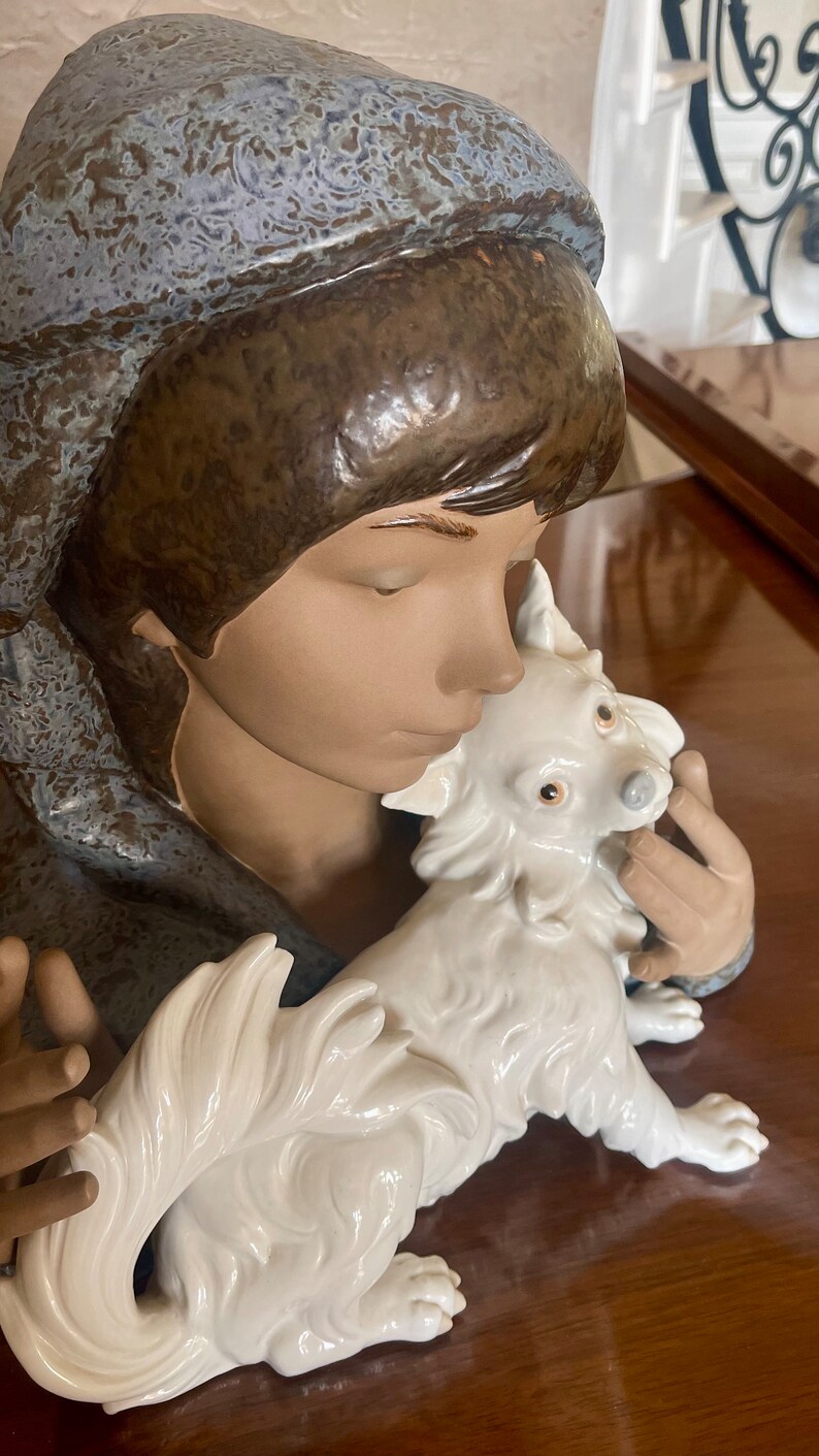 May include: A porcelain figurine of a girl with a blue and brown hooded cloak holding a white dog with a fluffy tail.