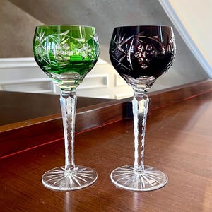 Puede incluir: Dos copas de vino de cristal tallado. Una tiene un cuenco verde, la otra un cuenco rojo oscuro. Ambas tienen tallos largos y transparentes y bases en forma de estrella. Las copas están sobre una superficie de madera oscura.