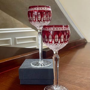May include: Two crystal wine glasses with ruby red bowls and clear stems. The glasses have a cut pattern design and sit on a wooden surface. A dark gray box is beneath the glasses.