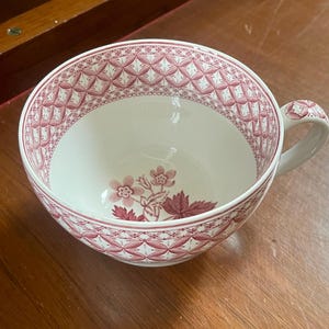 Puede incluir: Una taza de té de cerámica color crema con un patrón floral rosa oscuro en la base y un borde geométrico de celosía en el interior. La taza incluye un asa curva con un diseño floral rosa a juego en la parte superior.