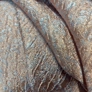 Könnte beinhalten: Nahaufnahme eines schimmernden Stoffes mit strukturierter Oberfläche. Der Stoff hat ein Muster aus verschlungenen Linien und einen Farbverlauf von hellbraun bis silber. Die Falten des Stoffes erzeugen Schatten und Glanzlichter.