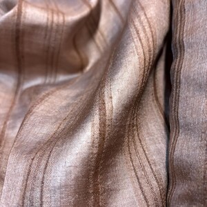 May include: Close-up of a shimmering fabric with a striped pattern. The fabric has a light brown base colour with darker brown vertical stripes. The material appears to be soft and flowing, with subtle variations in tone and texture.