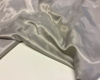 May include: A close-up of a silver satin fabric with a subtle embossed pattern of leaves. The fabric is draped and folded, showcasing its smooth texture and sheen.