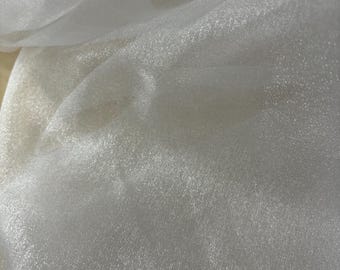 May include: Close-up of a flowing, off-white fabric with a subtle sheen. The material appears lightweight and slightly transparent, with soft folds and a delicate texture. The fabric's surface reflects light, creating highlights and shadows.